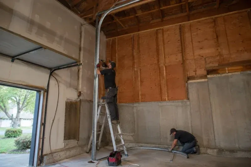 One person on a stepladder working beside a tall curved garage-door track while another kneels beside loose track sections in an unfinished garage. — High-Lift Garage Door Conversion, Wasaga Beach
